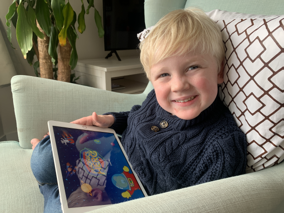 a three-year-old boy with blond hair sits in a wingbacked chair holding an iPad and smiling at the viewer